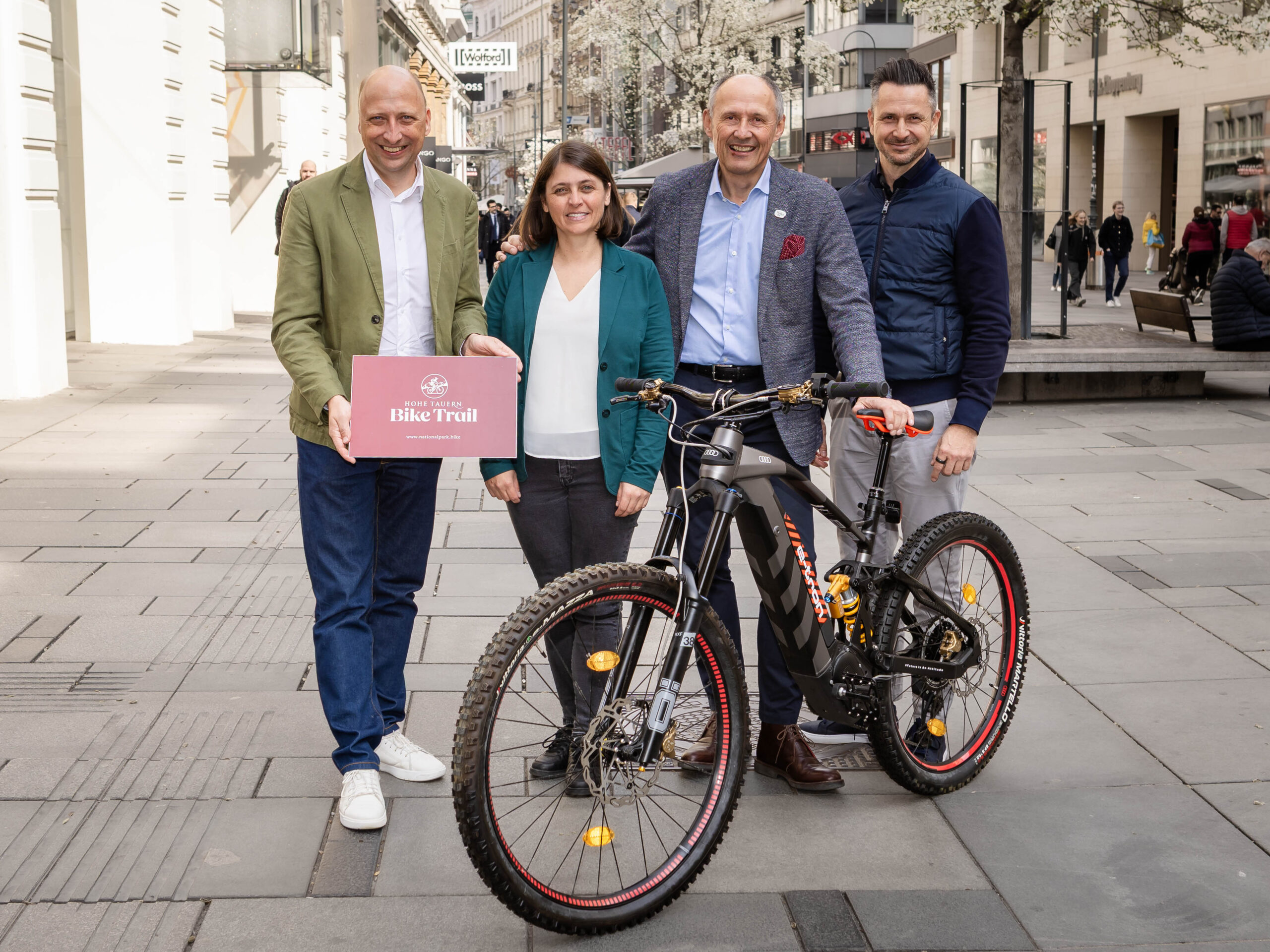 Vier Personen stehen mit einem Mountainbike auf einer Stadtstraße; eine hält ein rosa Bike Trail-Schild.