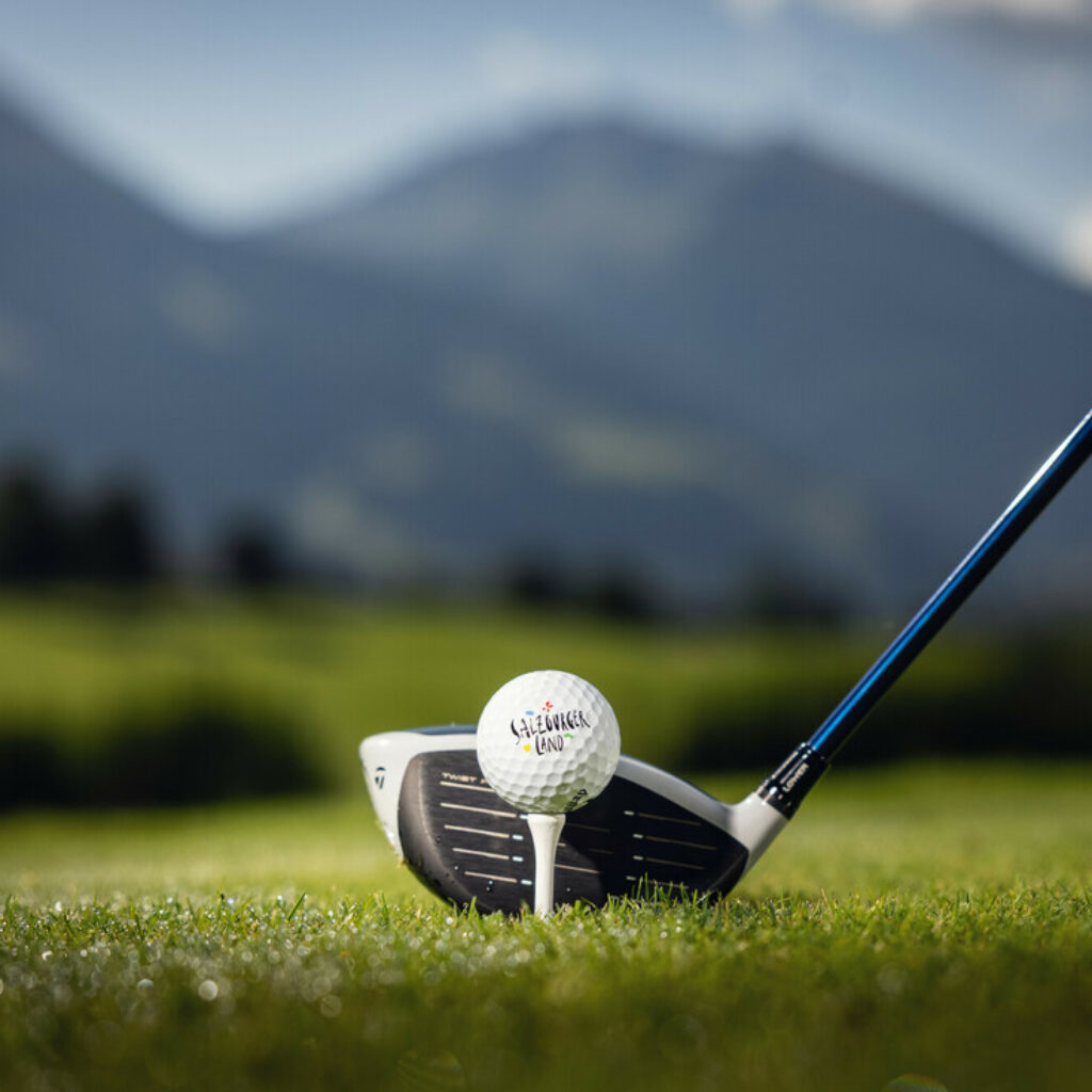Ein Golfschläger, der so positioniert ist, dass er einen Golfball auf einem grasbewachsenen Platz mit Bergen im Hintergrund schlägt.