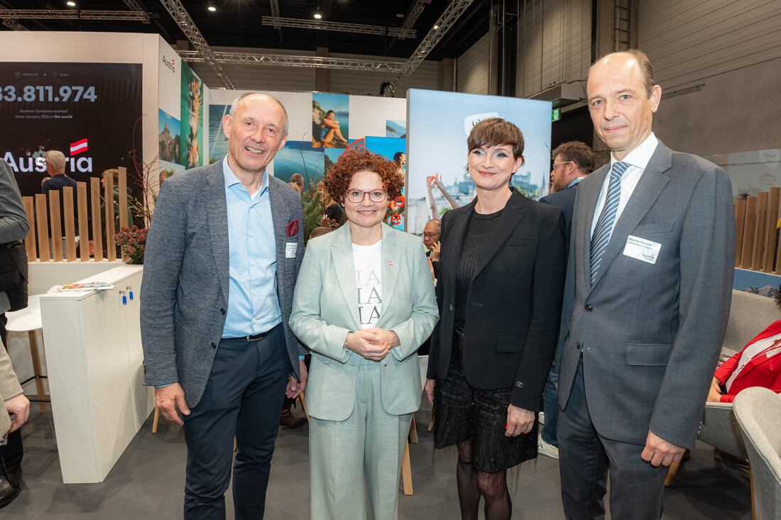 Vier Personen in Geschäftskleidung stehen zusammen und lächeln bei einer Veranstaltung oder Konferenz in einem Gebäude.