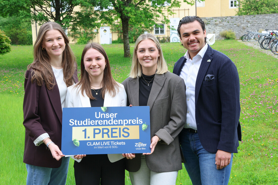 Vier lächelnde Menschen stehen draußen und halten ein Schild mit der Aufschrift Unser Studierendenpreis 1. Preis und einem anderen Text.