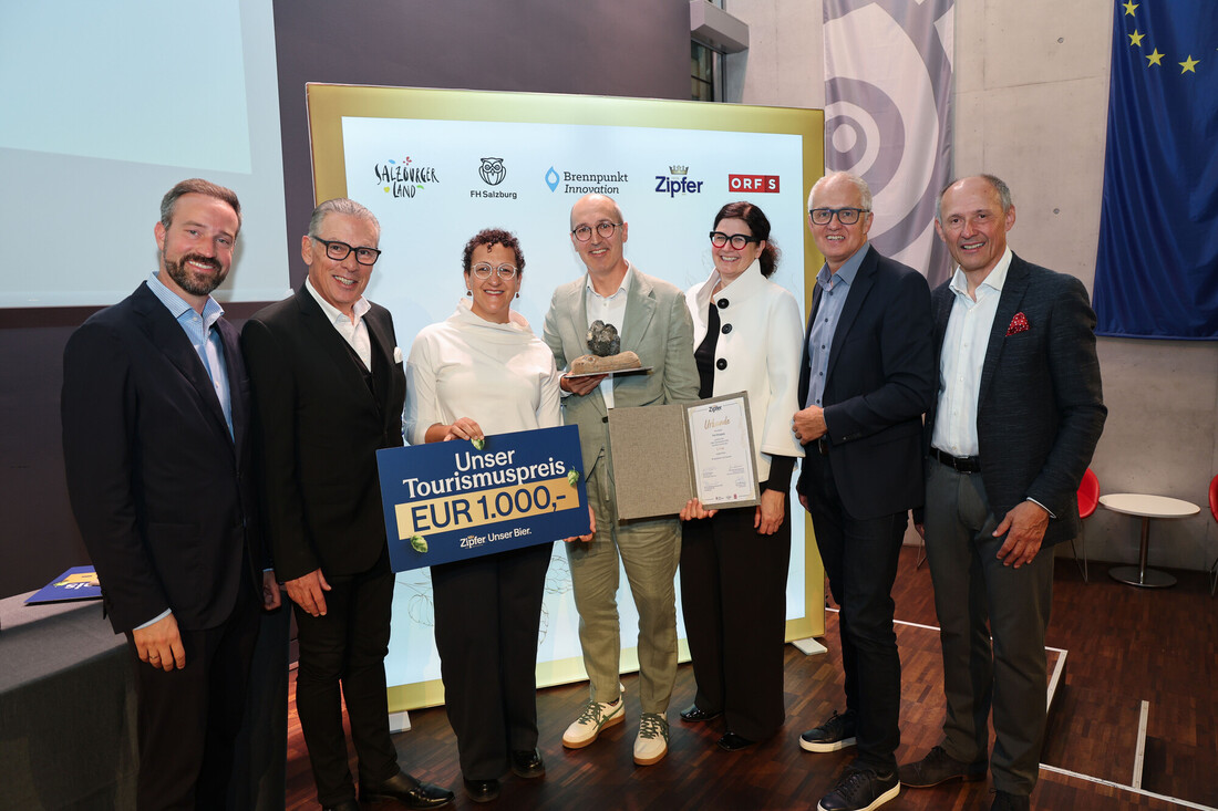 Sieben Personen stehen lächelnd in einem Raum zusammen, eine Person hält eine Trophäe, eine andere eine Urkunde in der Hand.