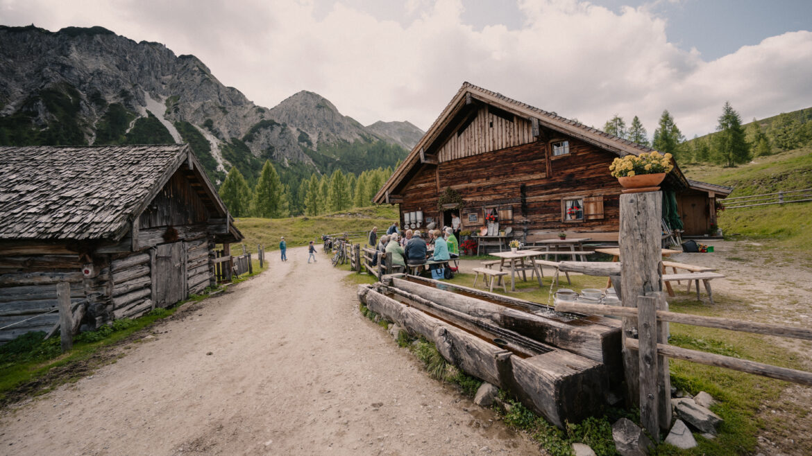 Menschen sitzen an Tischen im Freien bei rustikalen Holzhütten in einem Bergtal mit Bäumen und bewölktem Himmel.