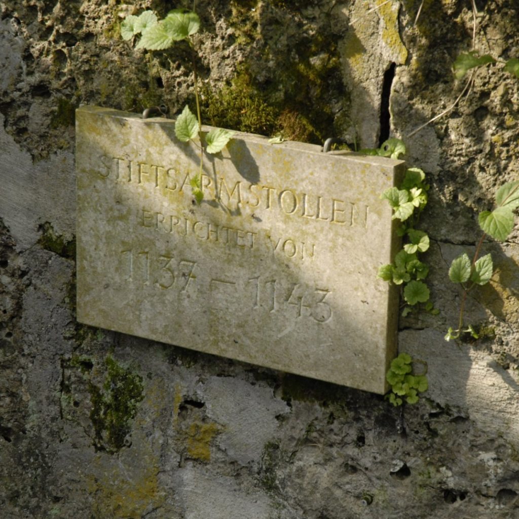 Eine verwitterte Steintafel mit eingraviertem Text und Daten, angebracht an einer moosbewachsenen, rissigen Mauer, die von grünen Ranken umrankt ist.