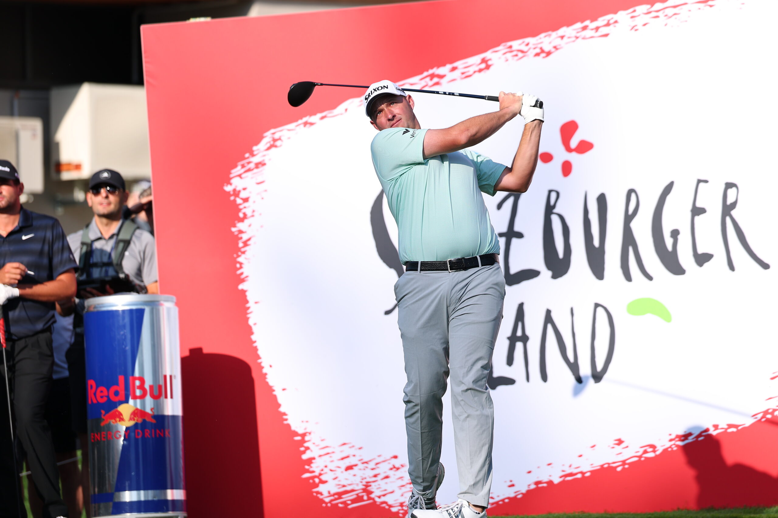 Ein Golfer schwingt einen Schläger auf einem Golfplatz, in dessen Nähe ein großes Cheeseburger Island-Schild und eine Red Bull-Dose stehen.