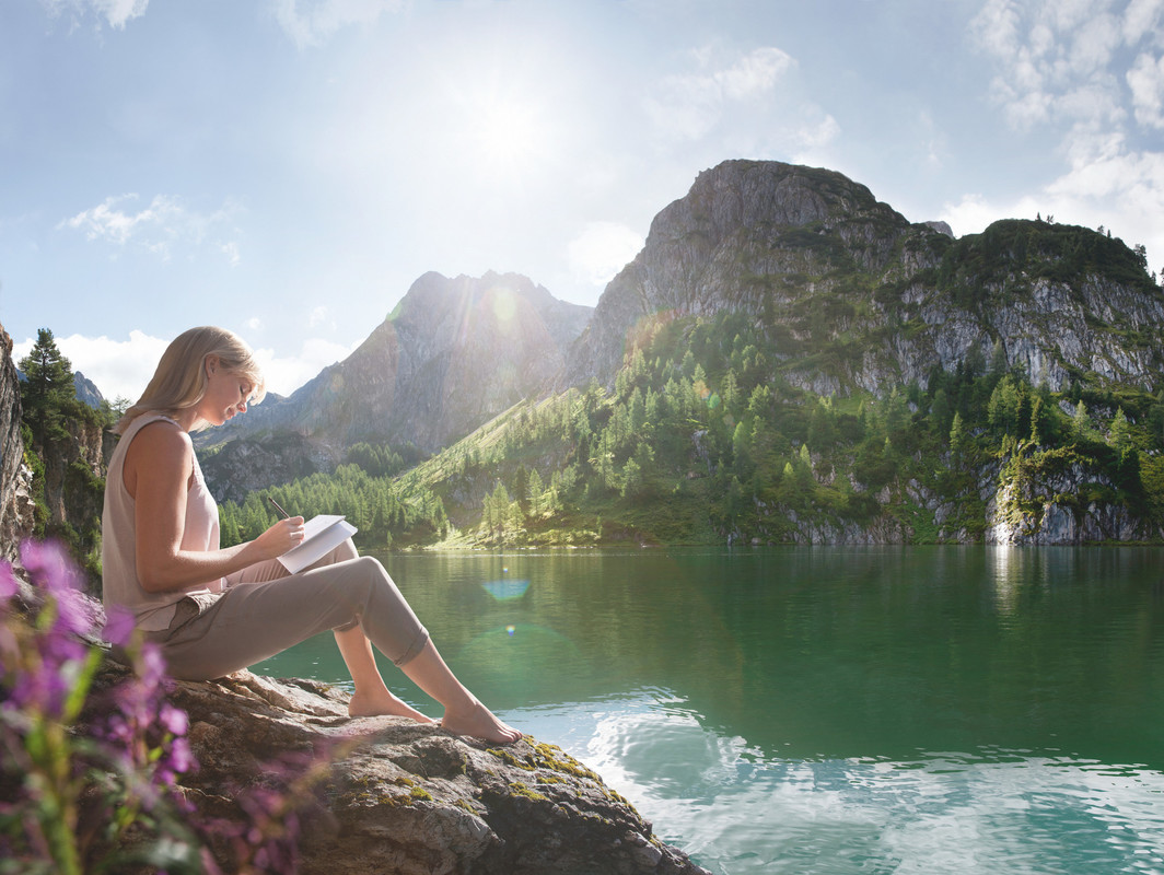Eine Frau, die auf einem Felsen am Seeufer ein Buch liest, im Hintergrund Berge und Bäume, unter strahlendem Sonnenschein.