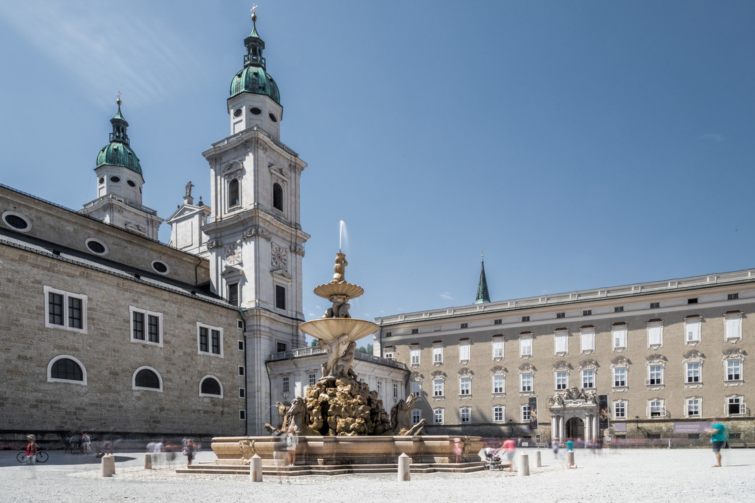 Ein historischer Platz mit einem großen, kunstvoll gestalteten Brunnen, umgeben von alten Steinhäusern und Kirchtürmen unter blauem Himmel.