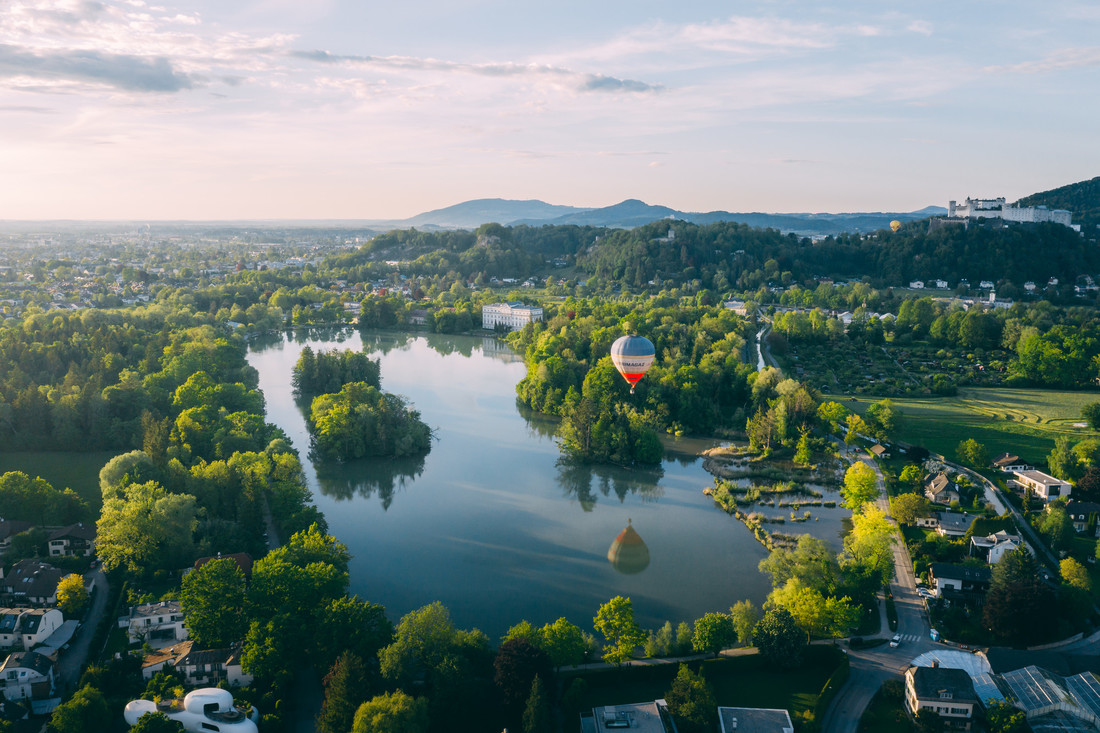 Ein Heißluftballon schwebt bei Sonnenuntergang unter klarem Himmel über einem See, der von Bäumen und Häusern umgeben ist.