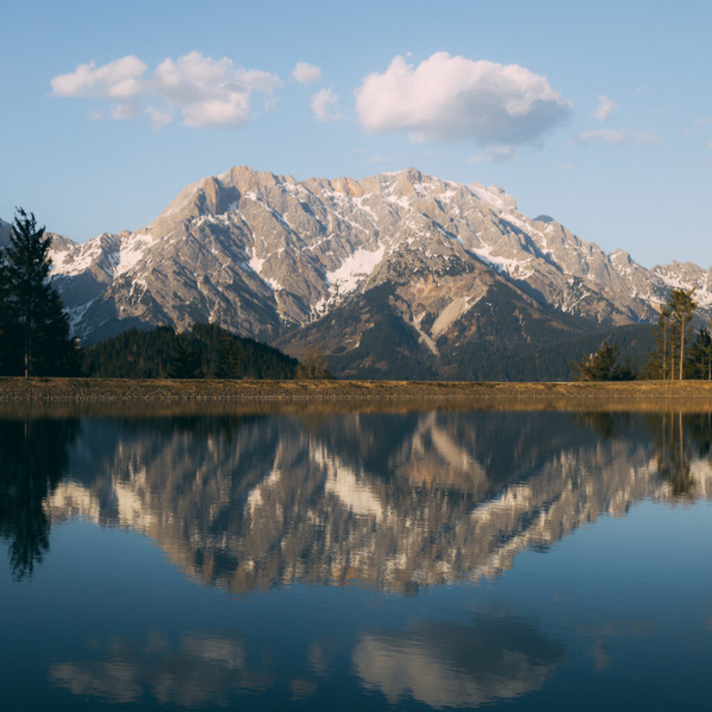 Ein schneebedeckter Berg spiegelt sich in einem ruhigen See unter einem klaren blauen Himmel mit wenigen Wolken.