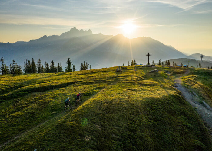 Zwei Radfahrer fahren bei Sonnenaufgang auf einem grasbewachsenen Hügel, mit Bergen und einem großen Kreuz im Hintergrund.
