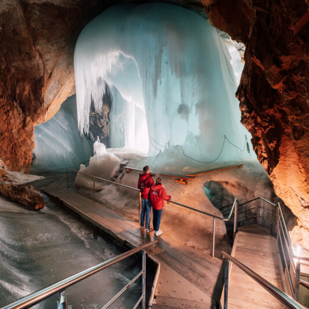 Zwei Menschen stehen auf einem Steg in einer Eishöhle mit großen Eiszapfen und vereisten Formationen.