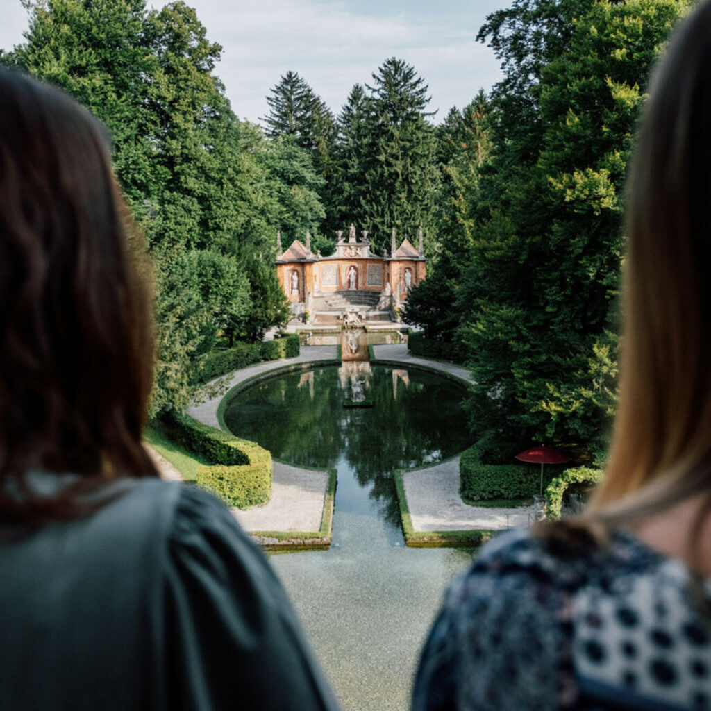Zwei Frauen blicken auf einen formalen Garten mit einem Teich und einem historischen Gebäude im Hintergrund.
