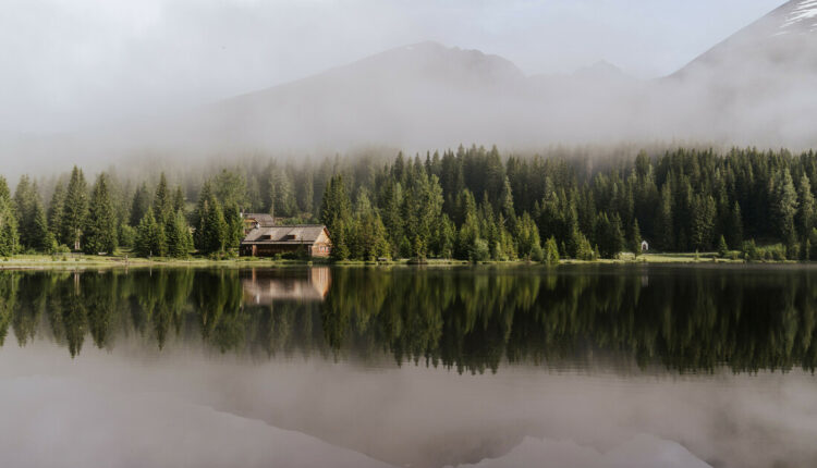 Eine Hütte an einem bewaldeten See, in dessen stillem Wasser sich Bäume und Berge spiegeln, während über dem See Nebel aufsteigt.