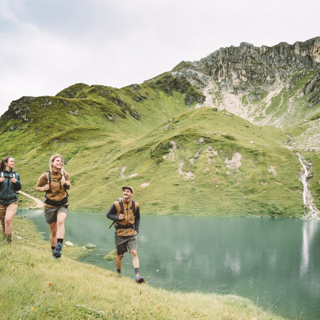 Drei Wanderer gehen an einem Bergsee entlang, umgeben von grünen Hügeln und felsigen Gipfeln unter einem bewölkten Himmel.
