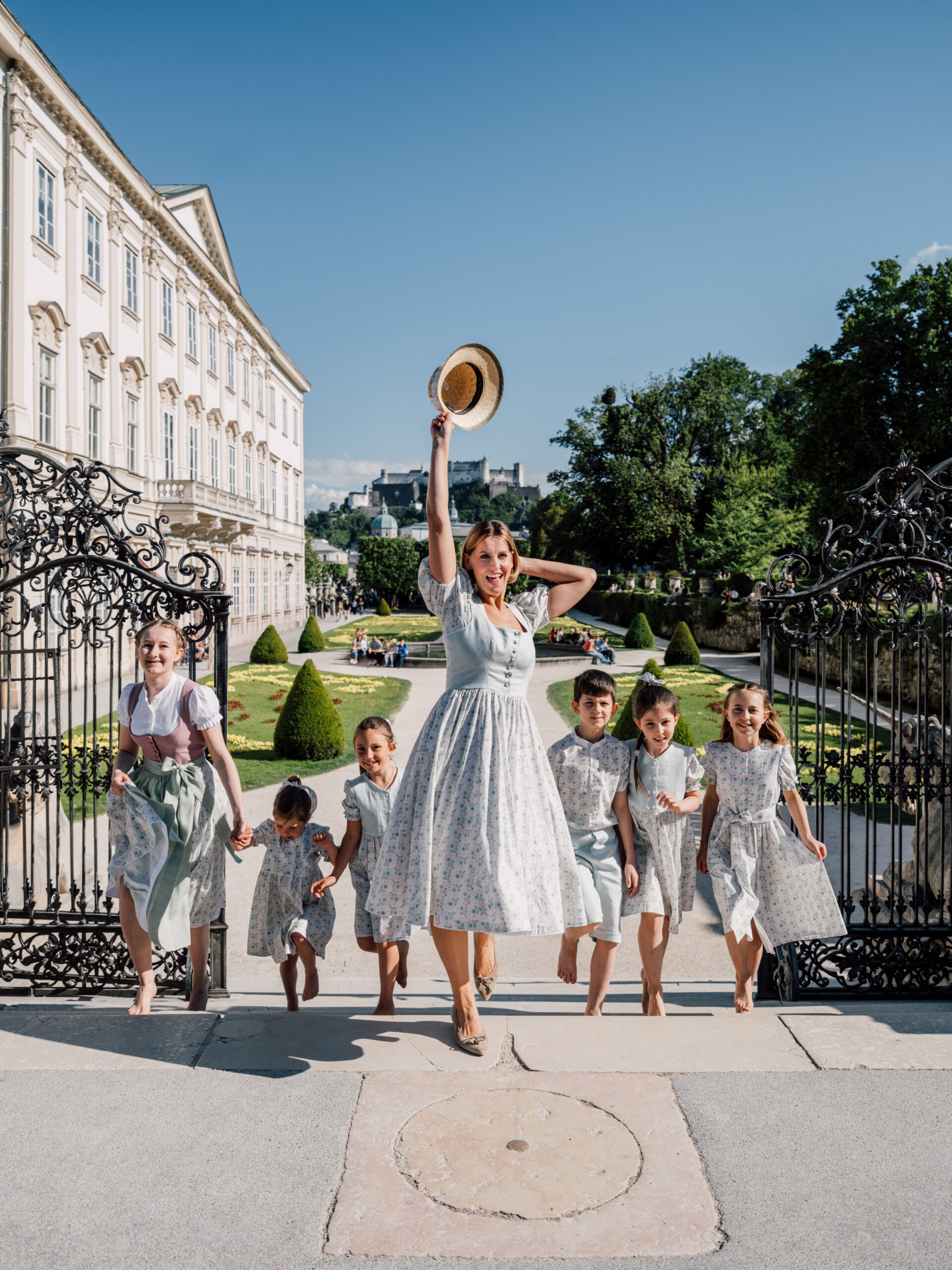 Eine Frau und sechs Kinder in einheitlichen Outfits schlendern an einem sonnigen Tag fröhlich durch kunstvoll verzierte Gartentore.