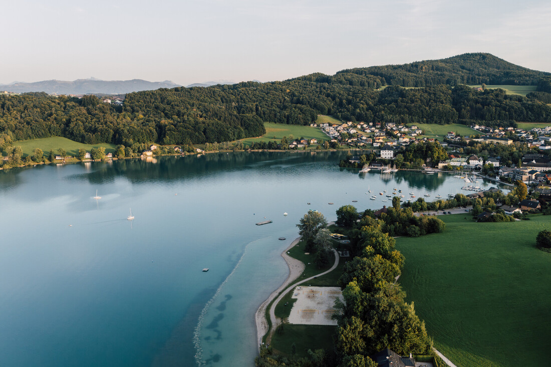 Luftaufnahme eines ruhigen Sees, umgeben von grünen Hügeln, Bäumen und einer kleinen Stadt mit vereinzelten Häusern.