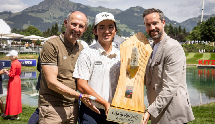 Drei lächelnde Männer mit einer großen Golftrophäe im Freien, Berge und See im Hintergrund.