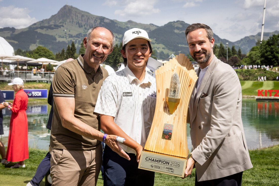 Drei lächelnde Männer mit einer großen Golftrophäe im Freien, Berge und See im Hintergrund.