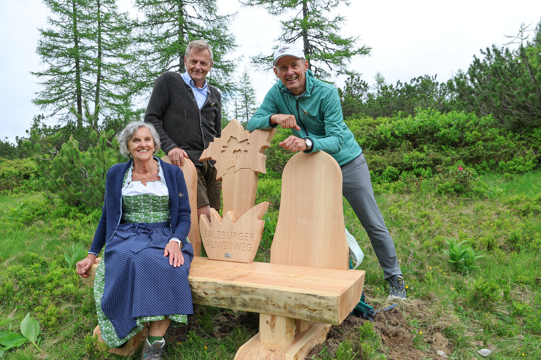 Drei lächelnde Menschen posieren neben einer Holzbank in einer malerischen Grünanlage mit Bäumen im Hintergrund.