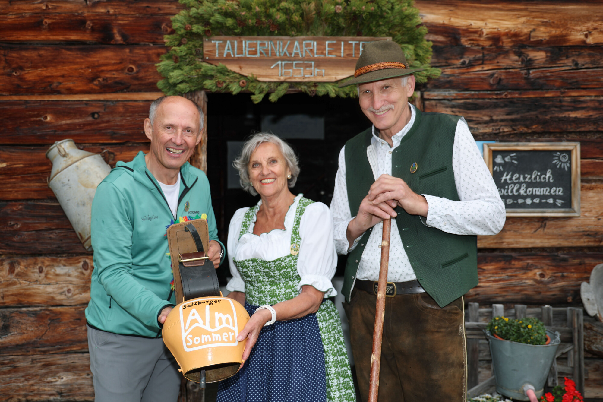 Drei lächelnde Menschen in traditioneller Alpenkleidung stehen vor einer rustikalen, geschmückten Holzhütte.