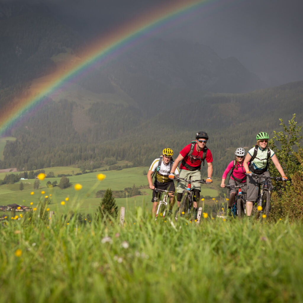 Vier Radfahrer fahren auf einem grasbewachsenen Weg bergauf, im Hintergrund sind ein Regenbogen und Berge zu sehen.