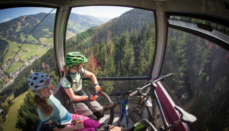 Zwei Mountainbiker mit Helmen fahren mit einer Seilbahn über ein bewaldetes Tal, die Fahrräder neben ihnen, mit schöner Aussicht.