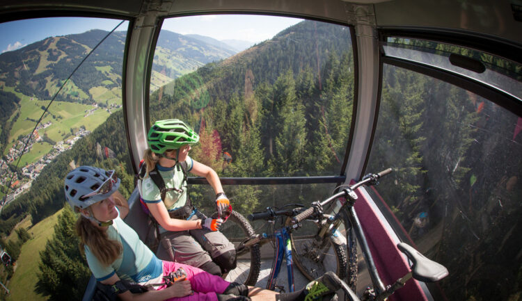 Zwei Mountainbiker mit Helmen fahren mit einer Seilbahn über ein bewaldetes Tal, die Fahrräder neben ihnen, mit schöner Aussicht.