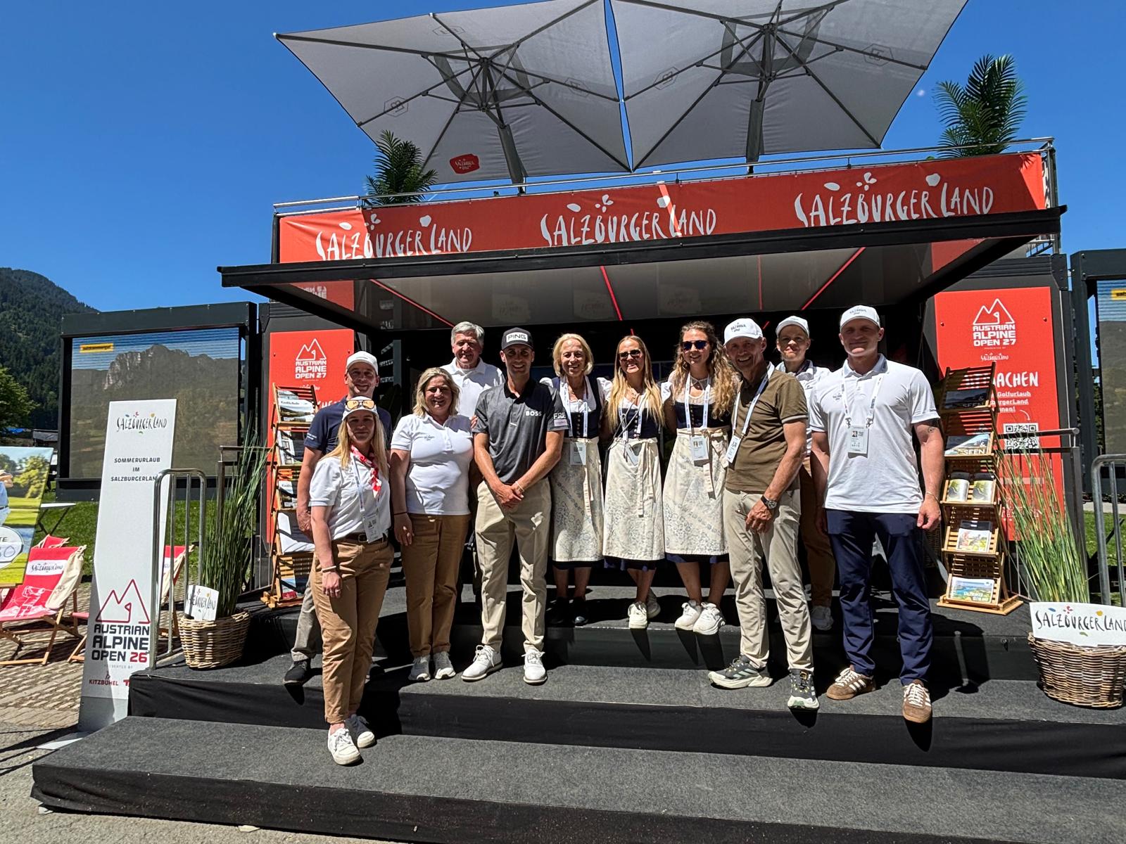 Eine Gruppe von Menschen steht an einem sonnigen Tag lächelnd auf den Stufen vor einem SalzburgerLand-Stand bei einer Freiluftveranstaltung.