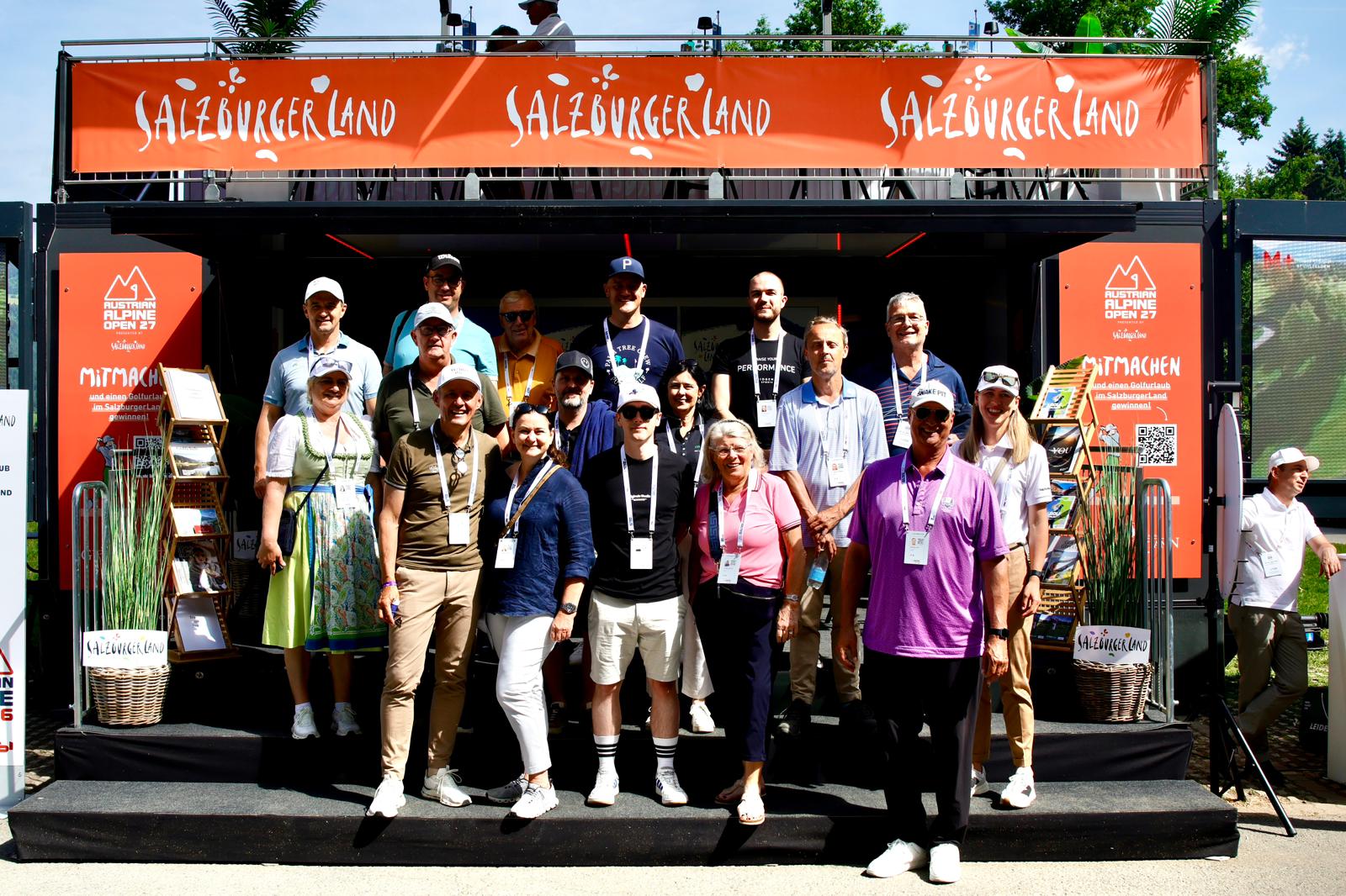 Eine Gruppe von Menschen posiert und lächelt an einem sonnigen Tag vor einem Salzburger Veranstaltungsstand.