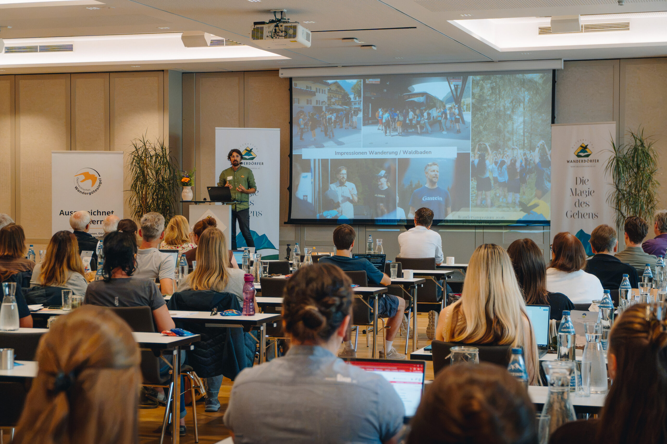 Ein Mann hält vor einem sitzenden Publikum in einem Konferenzraum, hinter ihm befindet sich eine große Leinwand, einen Vortrag.