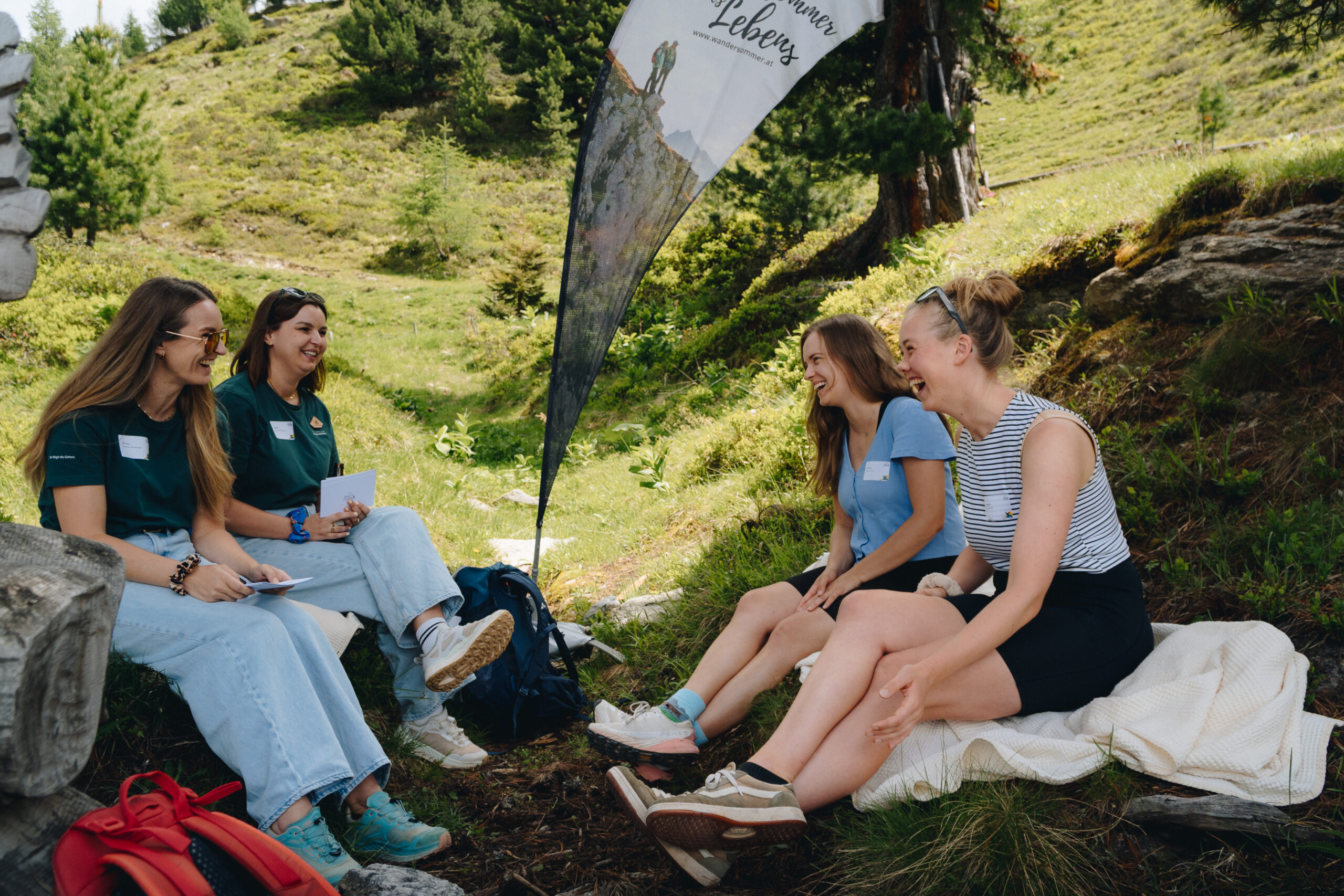 Vier junge Frauen sitzen gemeinsam im Freien auf einem grasbewachsenen Hügel, umgeben von Bäumen und einem Banner in der Nähe, und lachen.