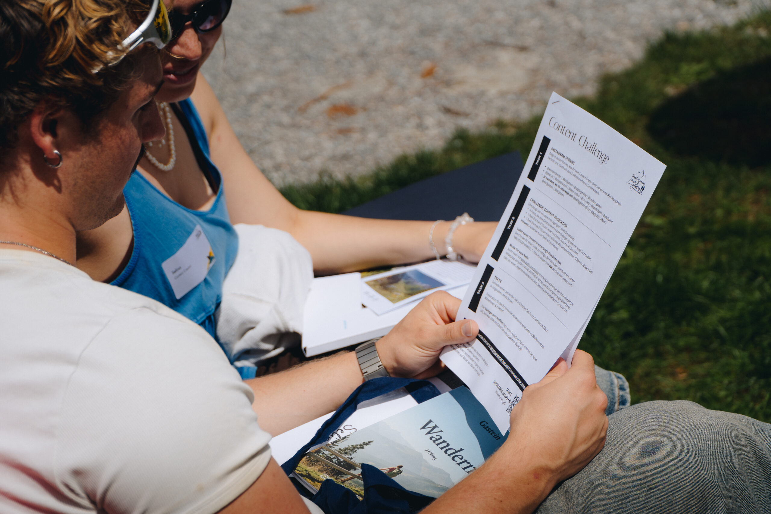Zwei Personen sitzen draußen, lesen ein gedrucktes Handout mit dem Titel „Contact Challenge“ und halten eine Zeitschrift in der Hand.