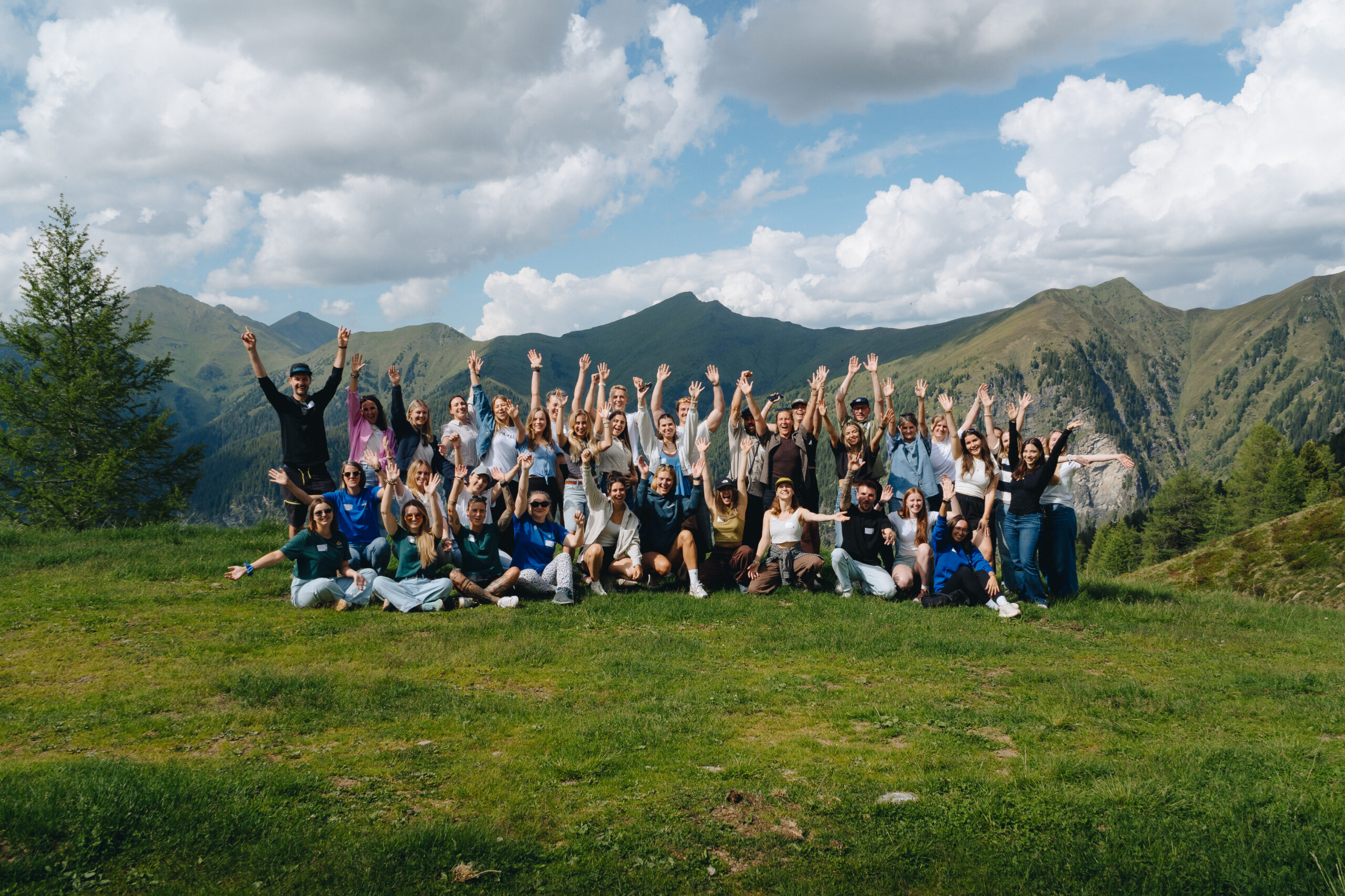 Eine große Gruppe von Menschen posiert mit erhobenen Händen auf einem grasbewachsenen Hügel, im Hintergrund sind Berge und Wolken zu sehen.