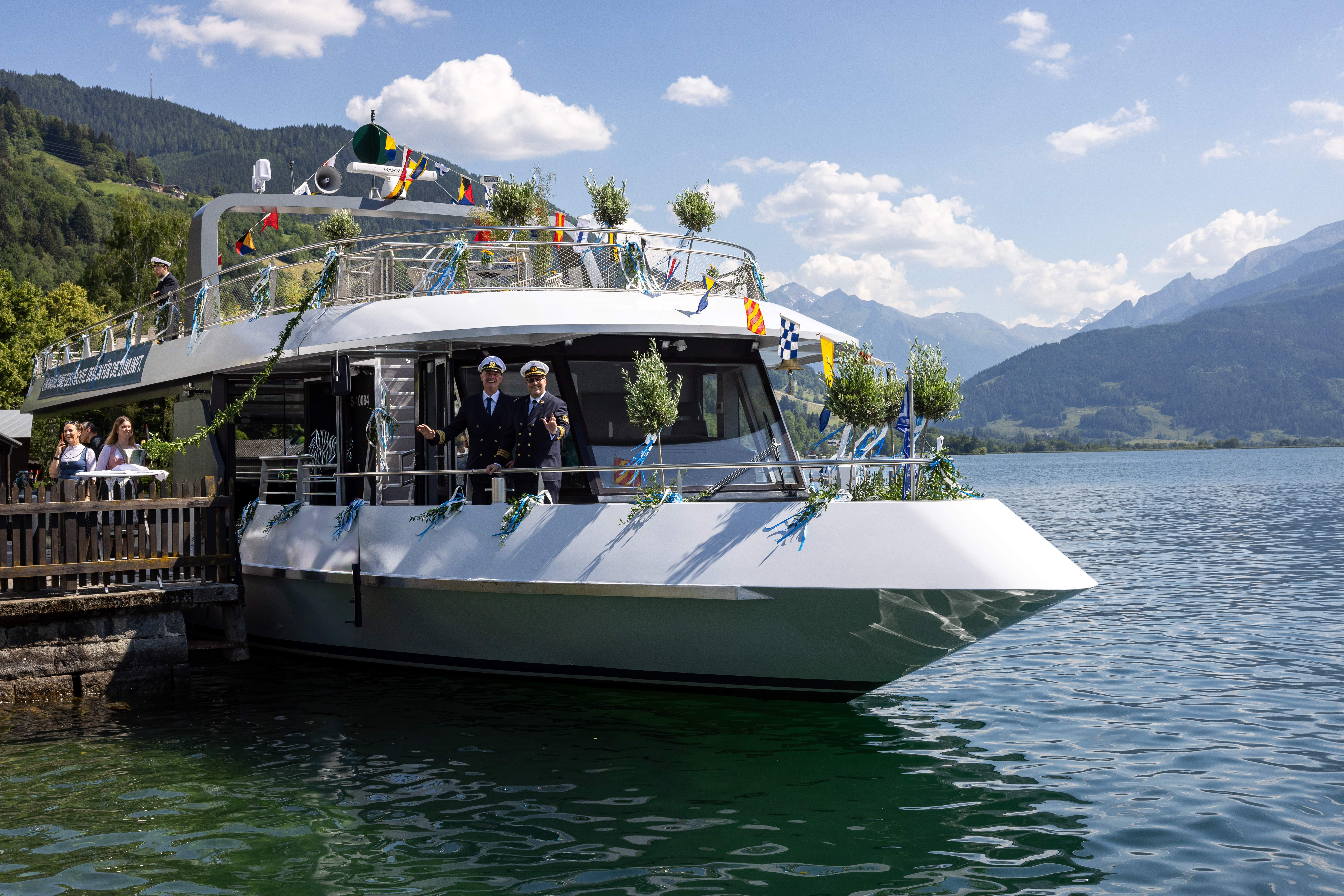 Ein geschmücktes weißes Boot mit Menschen in Uniform lag an einem malerischen See vor Anker, im Hintergrund waren Berge zu sehen.