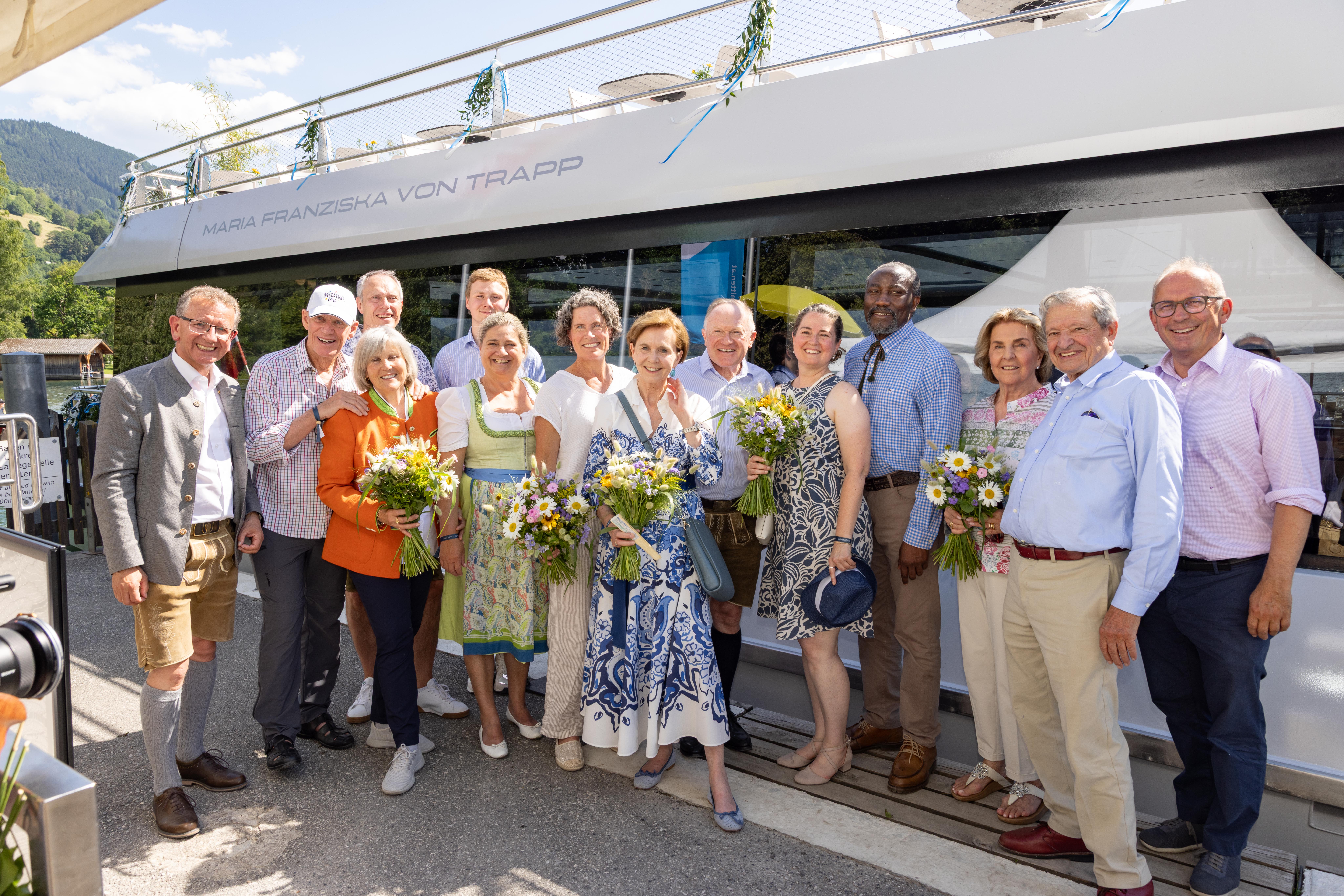 Eine Gruppe von Menschen, die lächeln und Blumen in den Händen halten, posiert vor einem Boot namens „Maria Franziska von Trapp“.