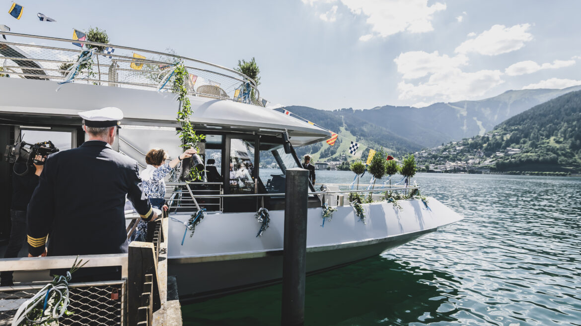 Menschen steigen an einem sonnigen Tag auf einem See, vor dessen Hintergrund sich Berge und eine Stadt abzeichnen, in ein geschmücktes Boot ein.