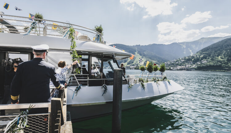 Menschen steigen an einem sonnigen Tag auf einem See, vor dessen Hintergrund sich Berge und eine Stadt abzeichnen, in ein geschmücktes Boot ein.