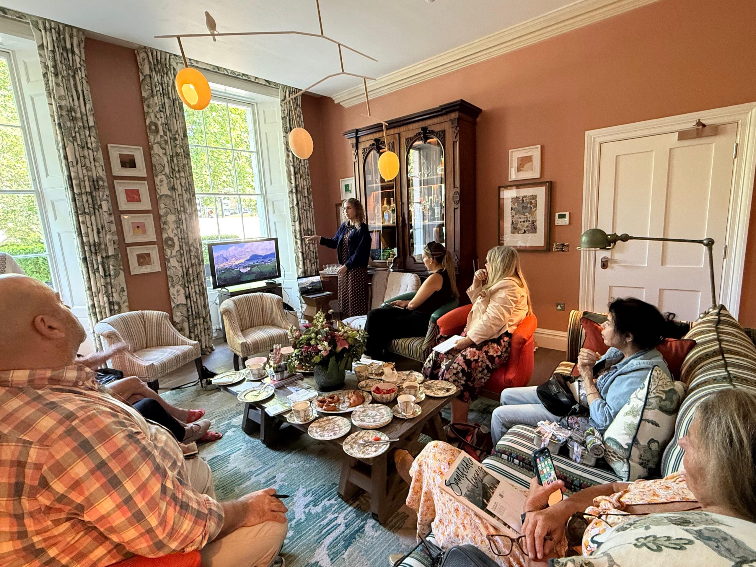 Eine Gruppe von Menschen hat sich in einem gemütlichen Wohnzimmer versammelt, wo es Tee und Snacks gibt, und hört einer Frau zu, die neben einem Fernseher steht.