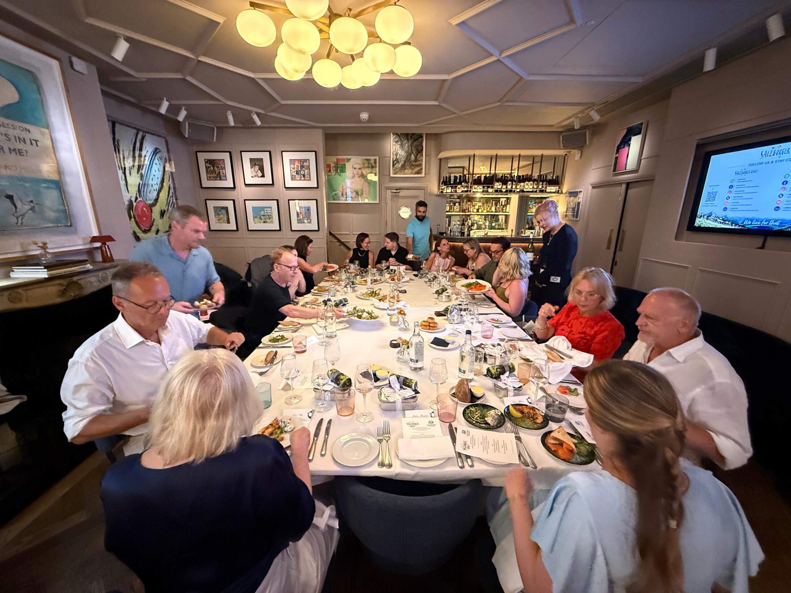 Eine Gruppe von Menschen sitzt gemeinsam an einem langen, elegant gedeckten Esstisch in einem warm beleuchteten Restaurant und genießt das Essen.