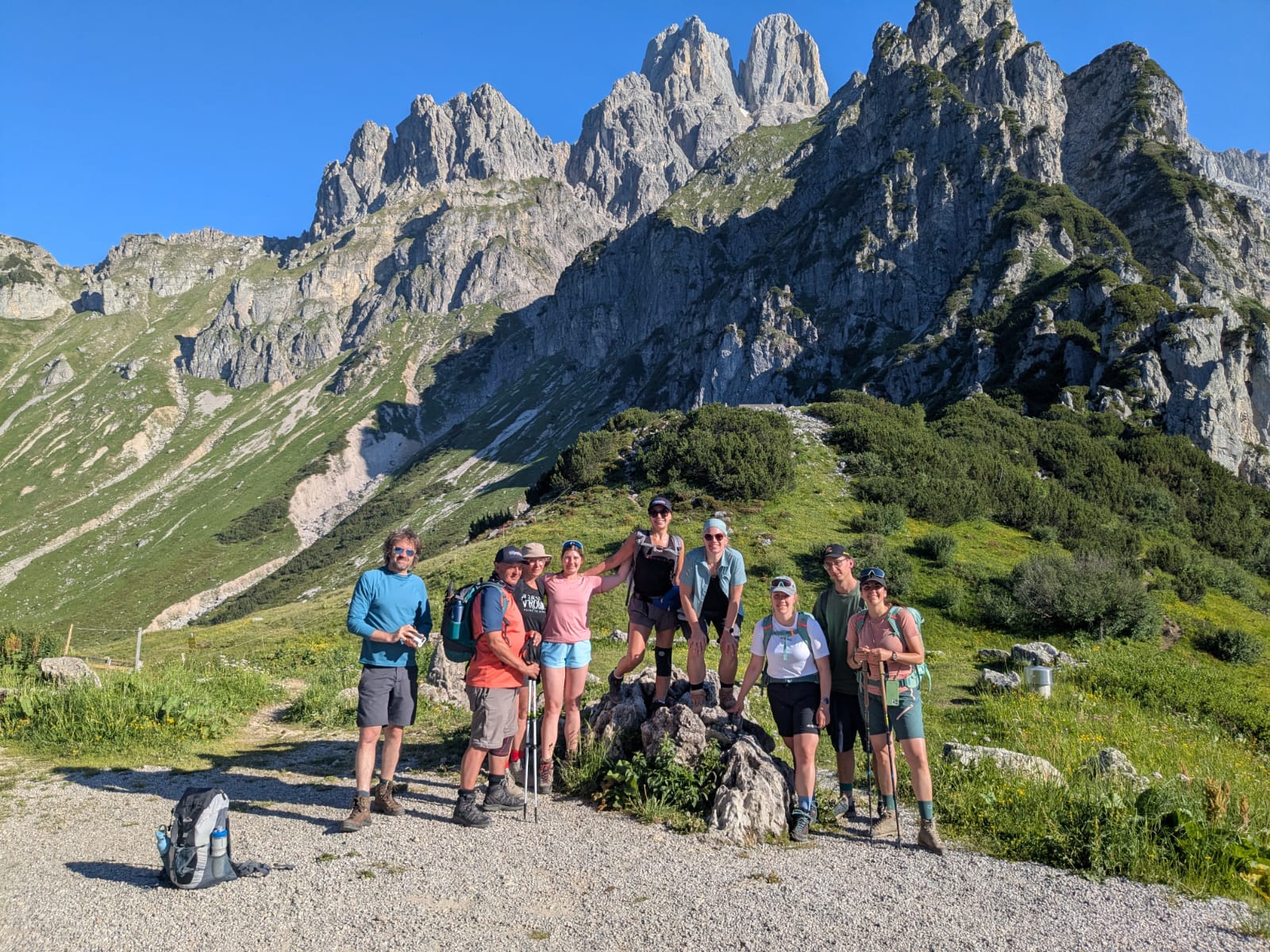 Eine Gruppe von Wanderern posiert auf einem felsigen Wanderweg, im Hintergrund sind hohe, schroffe Berge und grüne Landschaft zu sehen.