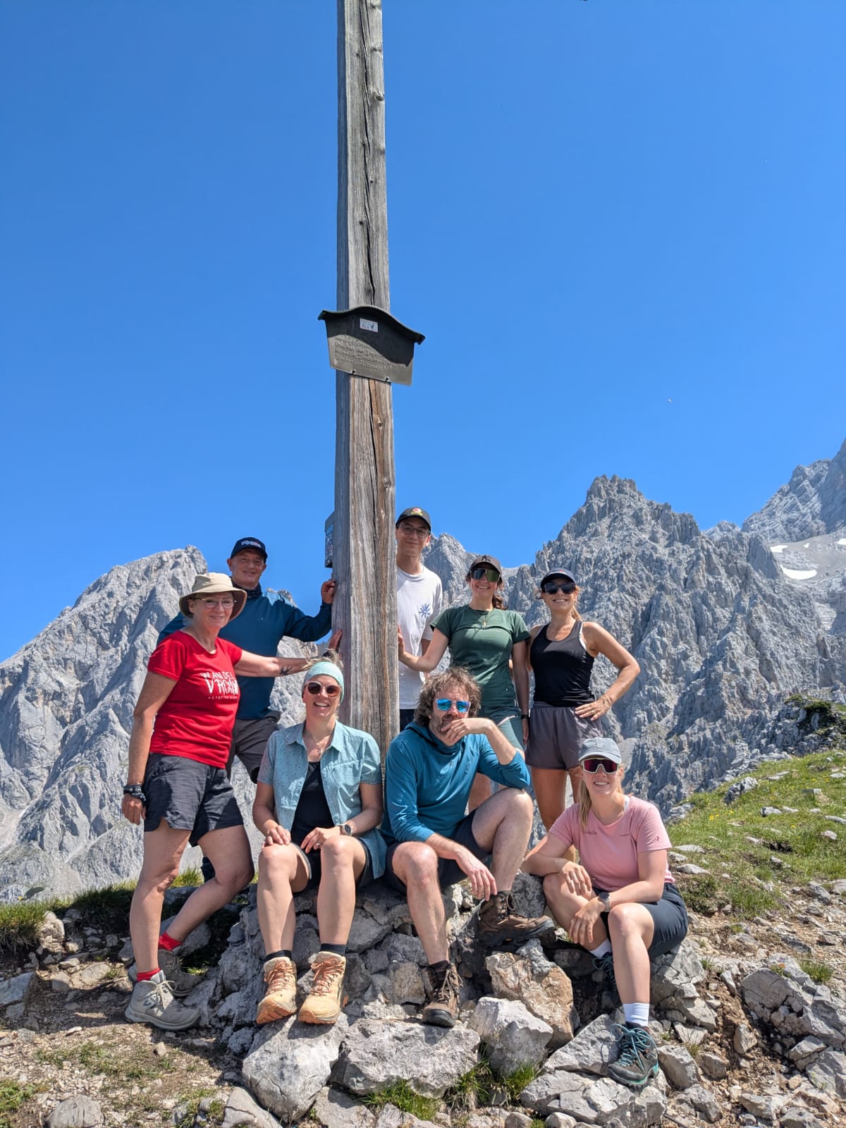Eine Gruppe von acht Wanderern posiert unter strahlend blauem Himmel neben einem Holzpfahl auf einem felsigen Berggipfel.