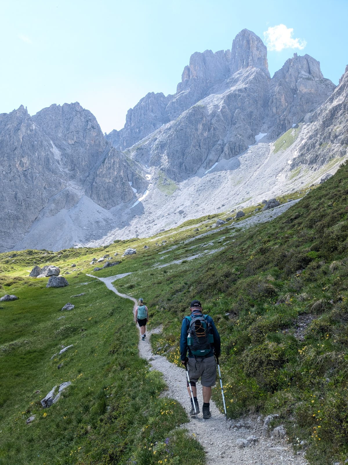 Zwei Wanderer gehen unter klarem Himmel auf einem Bergpfad entlang, der von felsigen Gipfeln und grünen Hängen gesäumt ist.