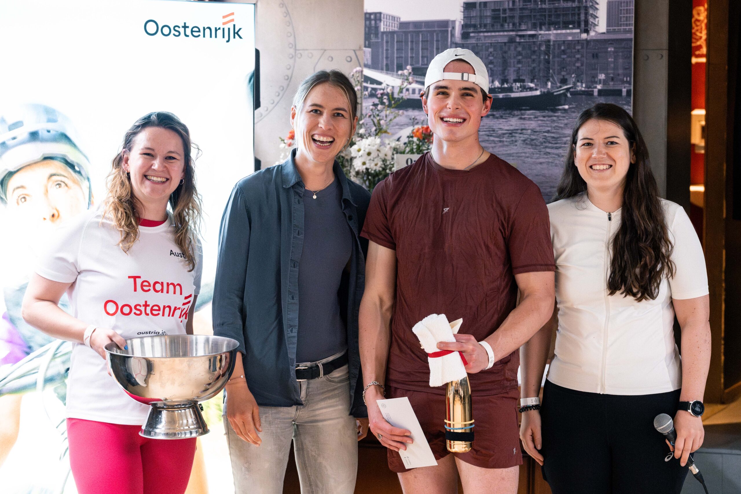 Vier lächelnde Personen posieren in einem Raum; eine hält einen Pokal, eine andere eine Flasche, im Hintergrund sind „Oostenrijk“-Logos zu sehen.
