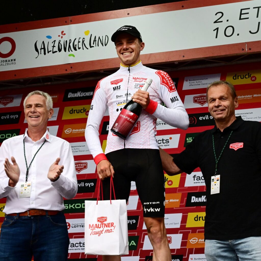 Radfahrer auf dem Podium, der eine Flasche und eine Geschenktüte in der Hand hält, lächelnd, neben ihm zwei Männer, im Hintergrund Veranstaltungsbanner.