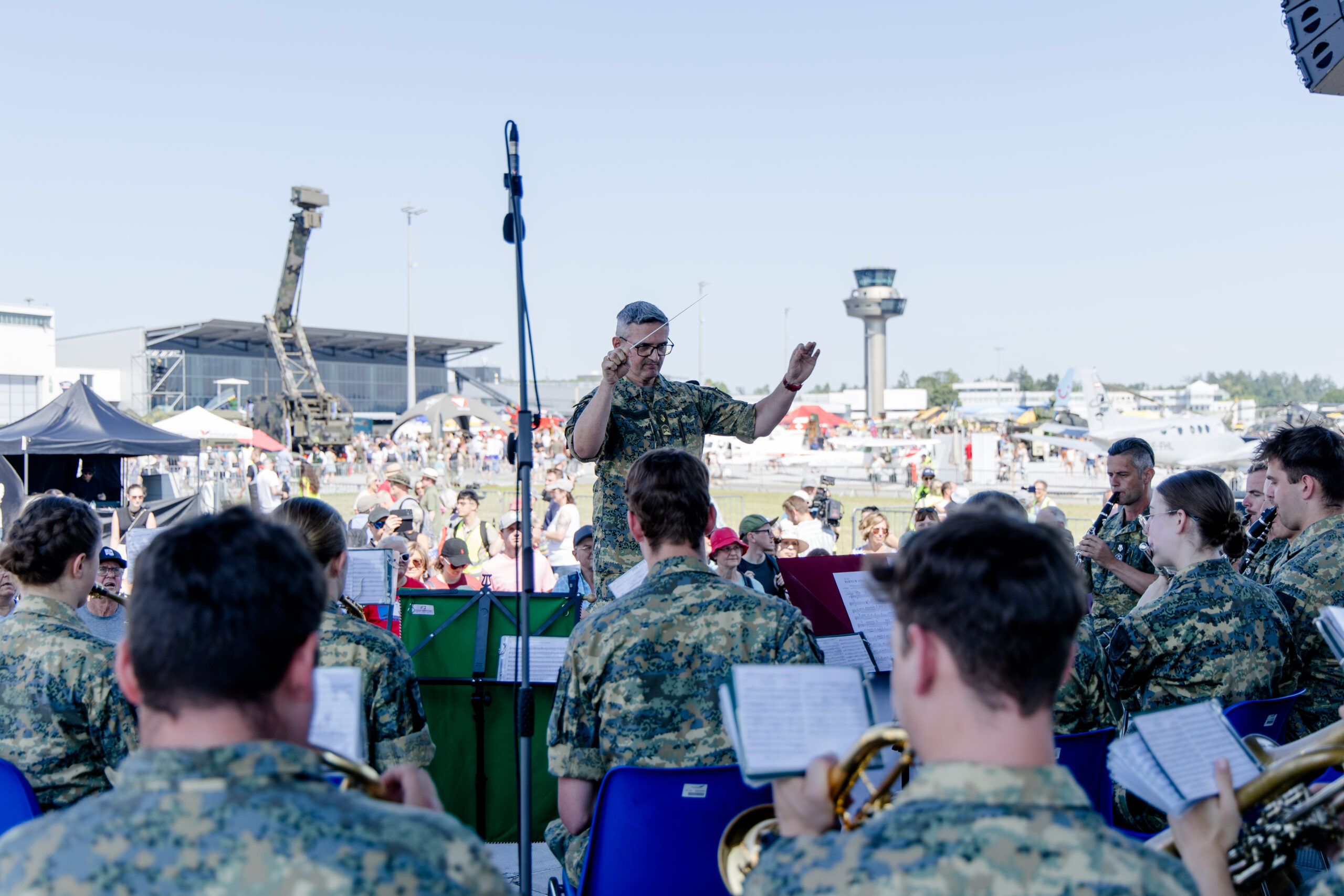 Eine Militärkapelle spielt unter freiem Himmel unter der Leitung eines Dirigenten, während eine Menschenmenge bei einer Veranstaltung auf einem Flugplatz zuschaut.