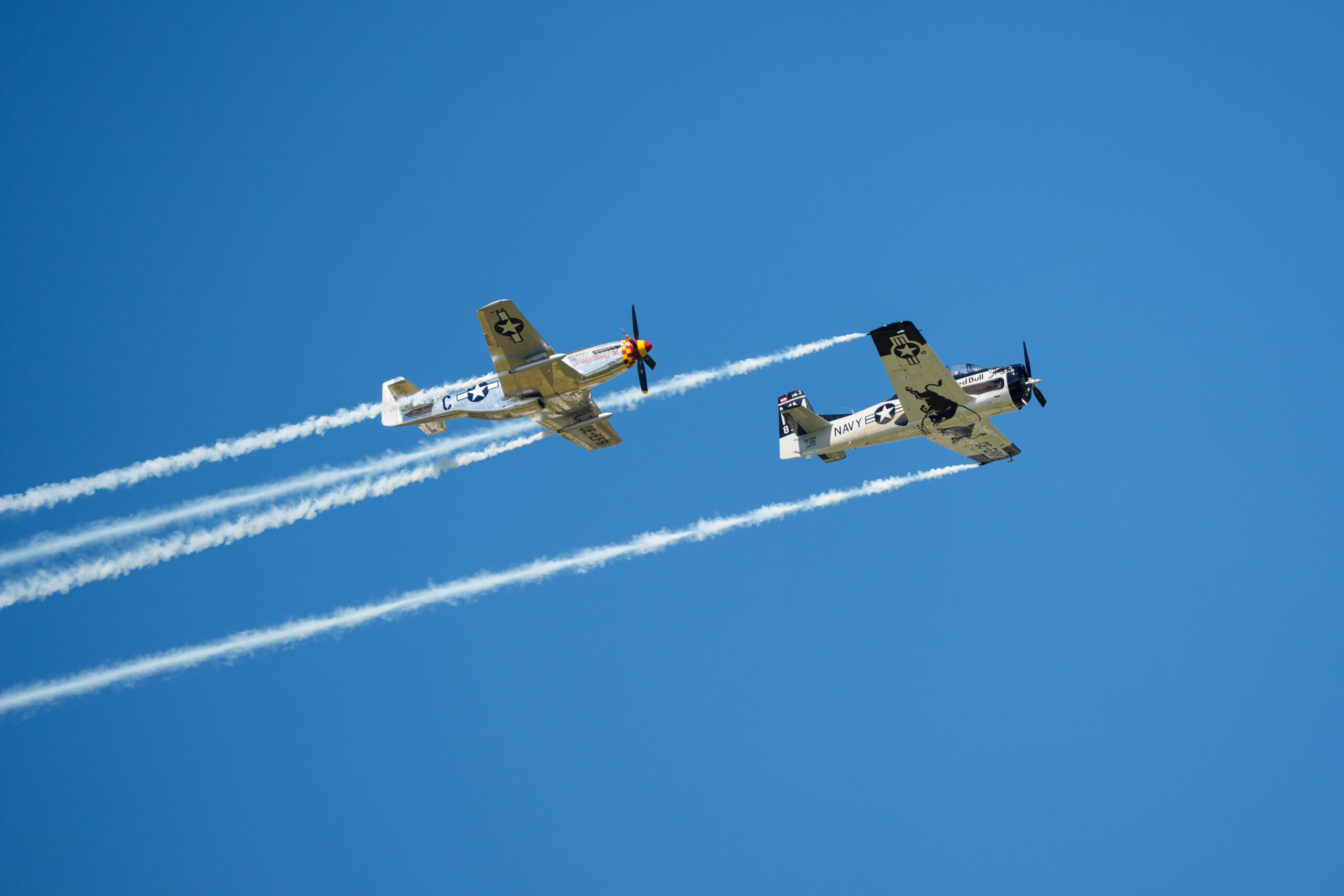 Zwei historische Militärflugzeuge fliegen nebeneinander und ziehen weiße Rauchspuren vor dem strahlend blauen Himmel.