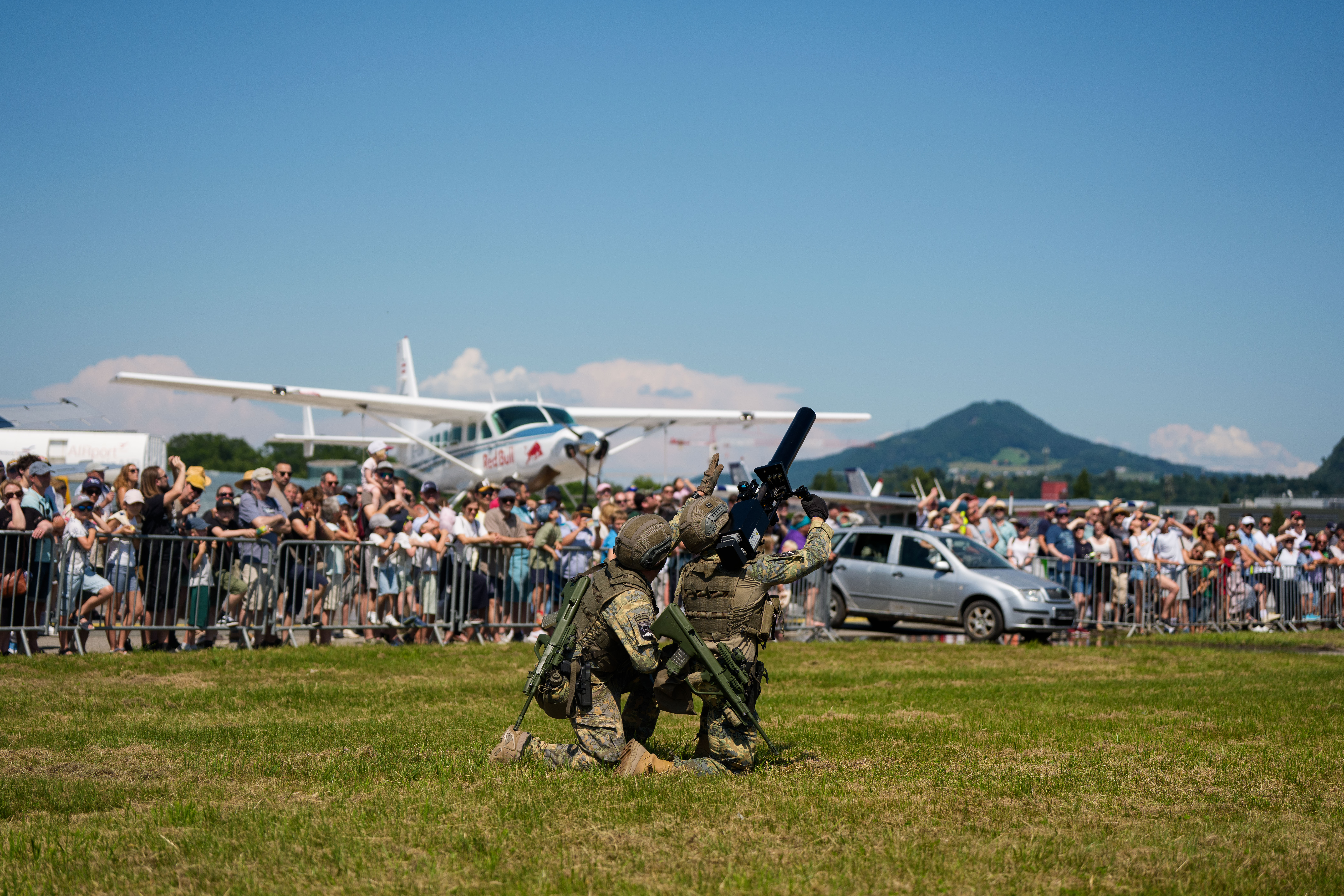 Drei Soldaten, die auf dem Rasen knien, treten bei einer Flugshow vor Publikum auf, im Hintergrund sind ein Flugzeug und Berge zu sehen.