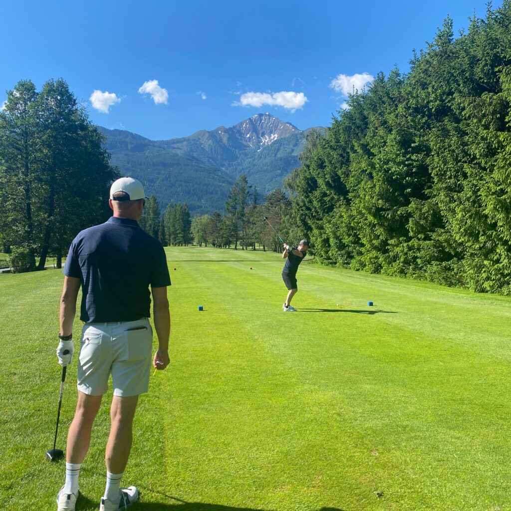 Zwei Menschen, die an einem sonnigen Tag Golf spielen, im Hintergrund Berge und Bäume.