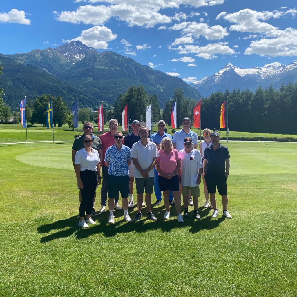 Eine Gruppe von Menschen, die an einem sonnigen Tag auf einem Golfplatz posiert, im Hintergrund sind Fahnen und Berge zu sehen.