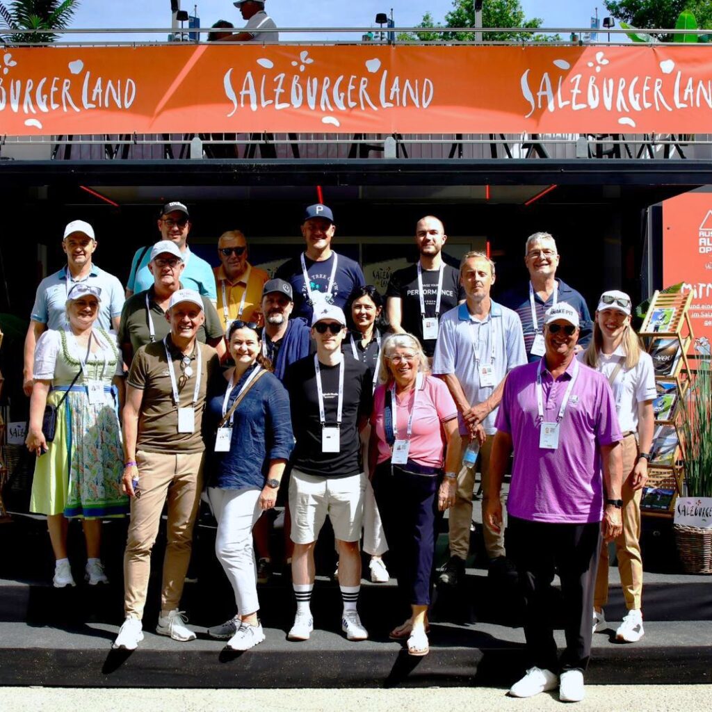 Eine Gruppe von Menschen posiert und lächelt vor einem Stand des SalzburgerLands bei einer Veranstaltung im Freien.