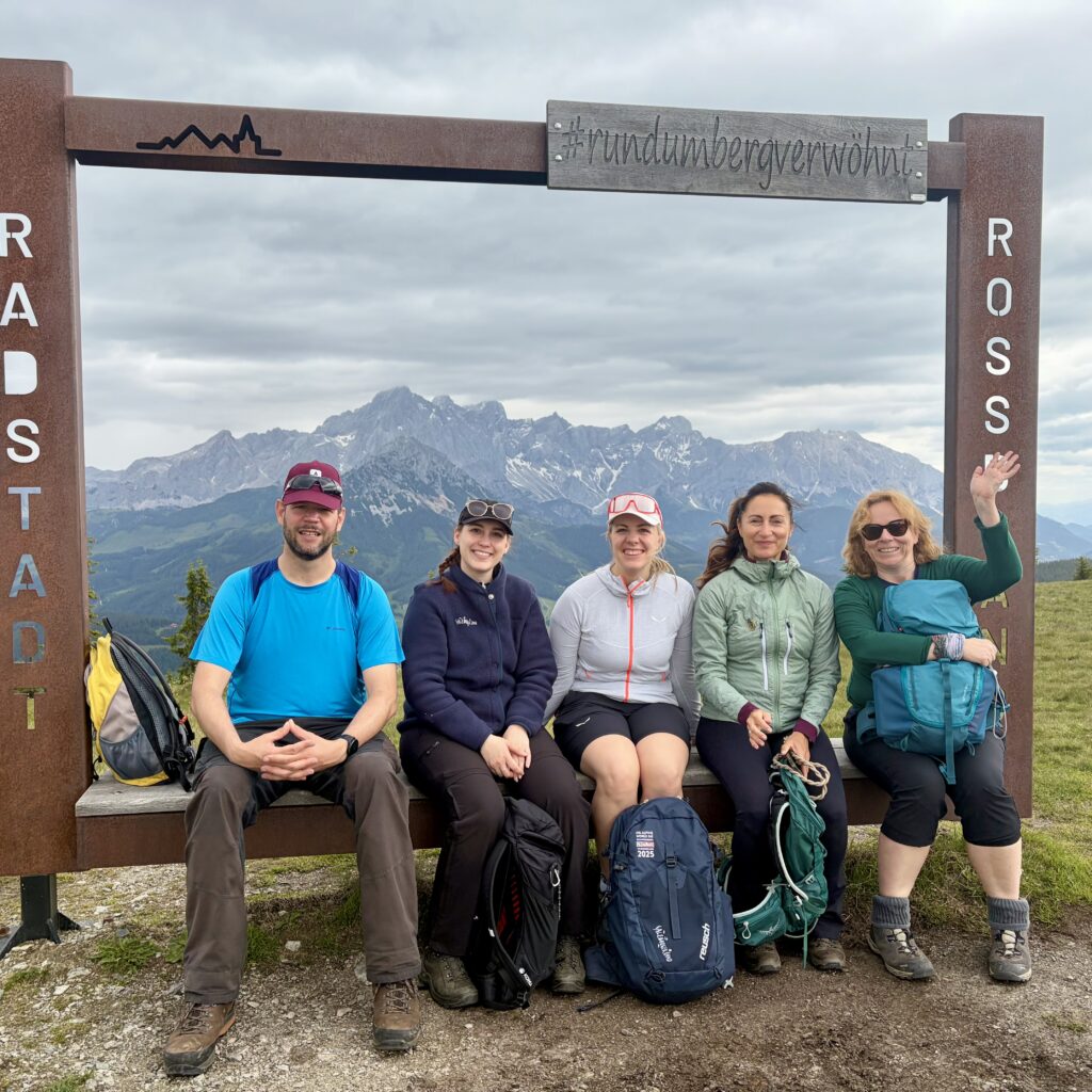 Fünf Wanderer sitzen lächelnd auf einer Bank vor dem Hintergrund von Bergen unter einem bewölkten Himmel.